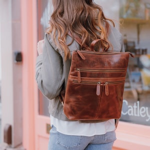 May include: A brown leather backpack with multiple zipped compartments. The backpack has a top handle and adjustable straps.