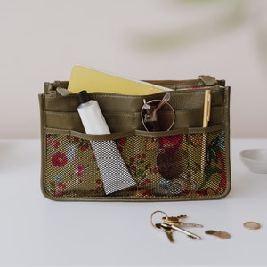 May include: A green zippered pouch with a floral pattern and mesh pockets. The pouch is filled with items such as a tube of lotion, sunglasses, a pen, and a small notebook.
