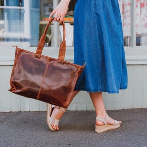 A large, brown leather tote bag with two handles is held by a woman walking on a sidewalk. The bag has a rectangular shape and a glossy finish. The model is wearing a blue denim skirt and beige sandals.