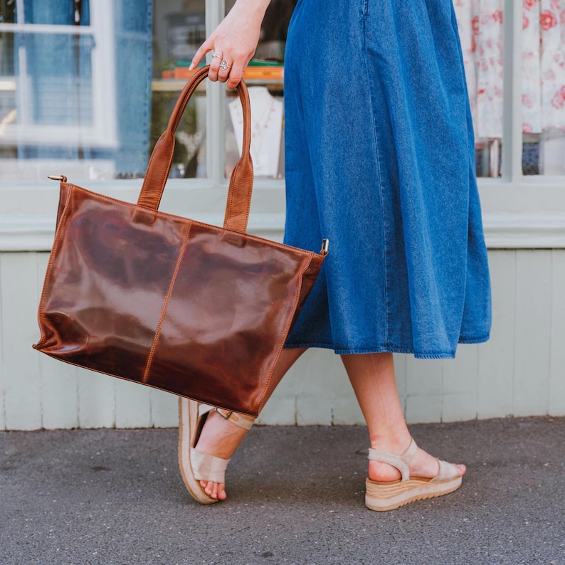 A large, brown leather tote bag with two handles is held by a woman walking on a sidewalk. The bag has a rectangular shape and a glossy finish. The model is wearing a blue denim skirt and beige sandals.