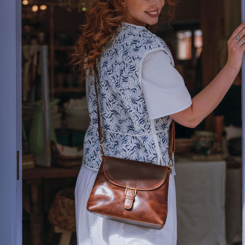 May include: A brown leather crossbody bag with a flap closure and a gold-colored buckle. The bag has a long strap and is worn over the shoulder. The bag is a rich, warm brown color.