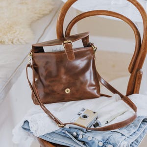 May include: A brown leather crossbody bag with a flap closure and gold-colored hardware sits on a wooden chair. A vintage camera and a tube of cream rest on the chair with a pair of blue jeans.