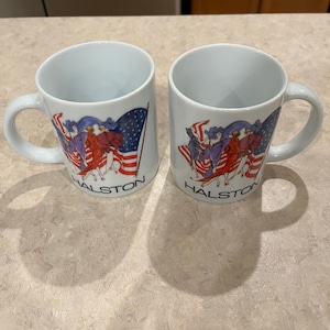 May include: Two white ceramic mugs with a patriotic design featuring an American flag and figures in red, white, and blue. The word "HALSTON" is printed below the design on each mug. The mugs have a curved handle.