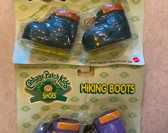 May include: Two pairs of Cabbage Patch Kids hiking boots in their original packaging. One pair is dark green with brown trim, the other is purple with orange trim. The packaging reads "Hiking Boots" and "Shoes" with the Cabbage Patch Kids logo.