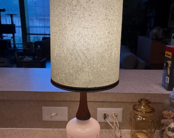 May include: A vintage table lamp with a cylindrical lampshade and a white, teardrop-shaped base. The base features a brown wood neck and a silver metal base. The lampshade is beige with a dark brown trim.