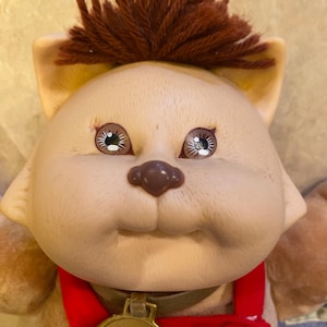 May include: A vintage Cabbage Patch Kids Koosas doll. The doll has a tan face with brown eyes, a brown nose, and a tuft of brown hair. It wears a red overall with a gold-colored medallion.