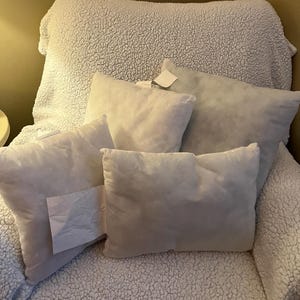 May include: A collection of white, rectangular pillows of various sizes, resting on a white, textured, sherpa-style chair. Some pillows have tags attached. The chair is in a room with a warm, neutral color scheme.