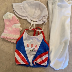 May include: A collection of children's clothing items, including a white sun hat, a pair of white sleeves, a pair of pink socks with white stars, and a red, white, and blue USA Olympics top.