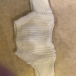 May include: A cream-colored, ribbed knit sweater with a turtleneck. The sweater appears to be made of a soft, possibly wool or cotton blend, and is folded. The ribbed texture is visible throughout the garment.