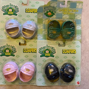 May include: Four pairs of Cabbage Patch Kids loafers in their original packaging. The shoes are in white, green, pink, and black. The packaging includes the text "LOAFER" and "SHOES". The shoes measure 14 inches.