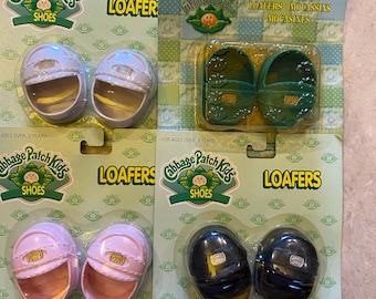 May include: Four pairs of Cabbage Patch Kids loafers in their original packaging. The shoes are in white, green, pink, and black. The packaging includes the text "LOAFER" and "SHOES". The shoes measure 14 inches.