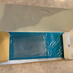 May include: An open cardboard box with a teal item inside, wrapped in clear plastic. The box is beige and has a curved handle on the top flap. The item is rectangular and appears to be a rug or mat.