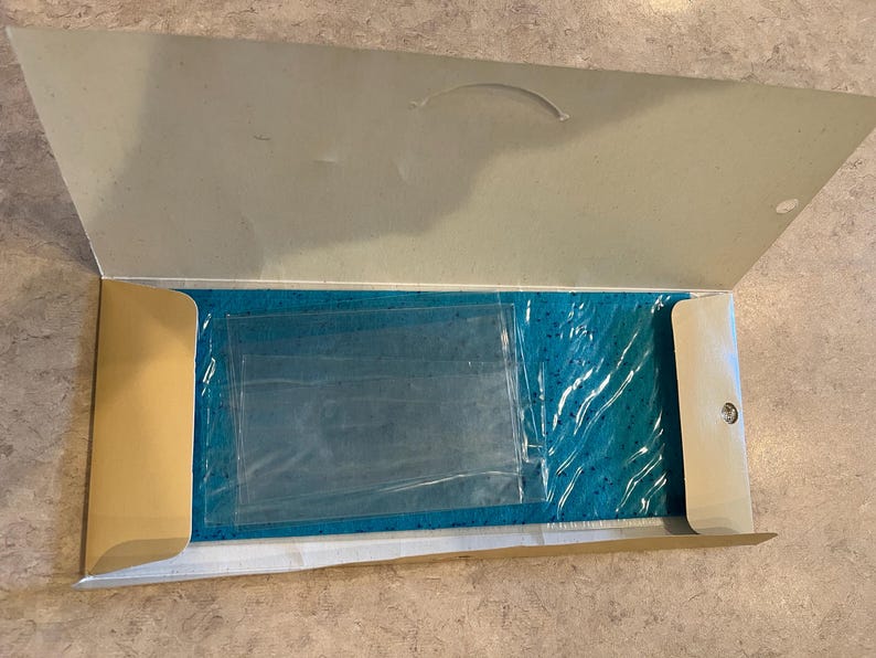 May include: An open cardboard box with a teal item inside, wrapped in clear plastic. The box is beige and has a curved handle on the top flap. The item is rectangular and appears to be a rug or mat.