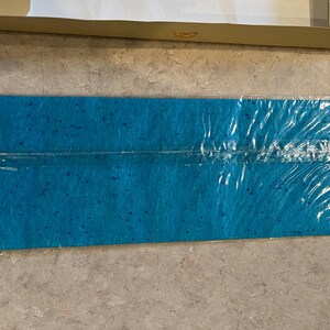 May include: A rectangular, turquoise felt sheet with darker blue speckles, sealed in clear plastic. The felt is positioned on a light-colored surface, with a portion of a cardboard box visible in the background.