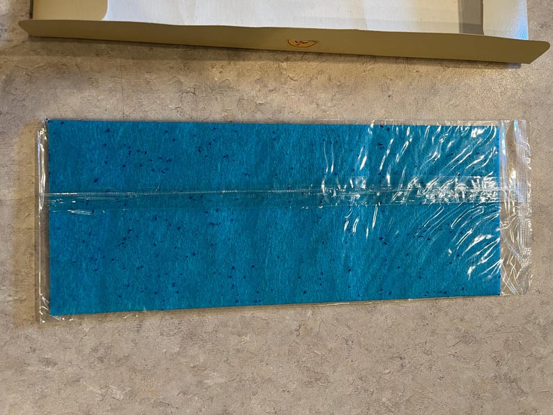 May include: A rectangular, turquoise felt sheet with darker blue speckles, sealed in clear plastic. The felt is positioned on a light-colored surface, with a portion of a cardboard box visible in the background.