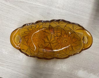 May include: Amber-colored, oval-shaped glass dish with a floral design. The dish features two handles and a scalloped edge. The floral pattern includes sunflowers and starbursts.
