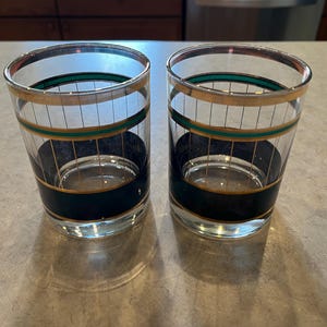 May include: Two clear drinking glasses with gold rims and accents. Each glass has a dark green and black band around the middle and vertical gold lines. The glasses are sitting on a light-colored surface.