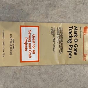 May include: A package of Dritz Mark B. Gone Tracing Paper. The package is tan with red and white text. The text reads "Great for All Sewing and Craft Projects." The package contains one 10 in x 11 3/8 in sheet.