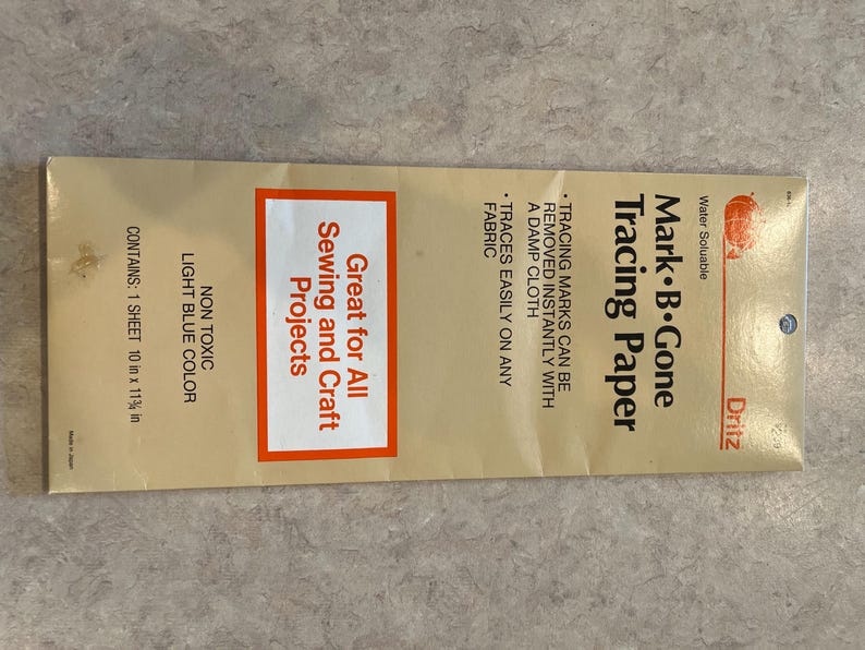 May include: A package of Dritz Mark B. Gone Tracing Paper. The package is tan with red and white text. The text reads "Great for All Sewing and Craft Projects." The package contains one 10 in x 11 3/8 in sheet.