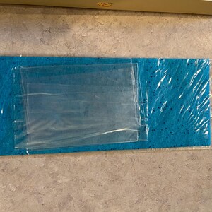 May include: A rectangular, turquoise cleaning cloth with a speckled pattern, packaged in clear plastic. The cloth is folded and has a clear plastic pouch on top. The packaging is sealed.
