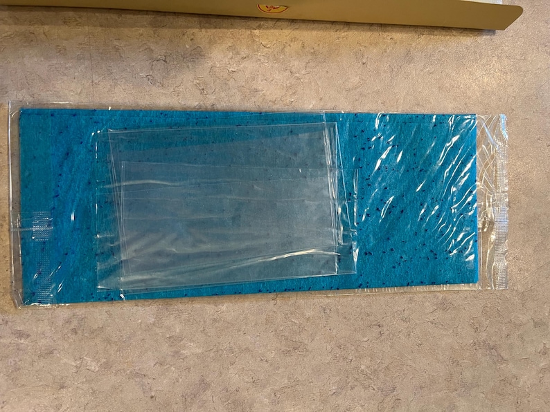 May include: A rectangular, turquoise cleaning cloth with a speckled pattern, packaged in clear plastic. The cloth is folded and has a clear plastic pouch on top. The packaging is sealed.
