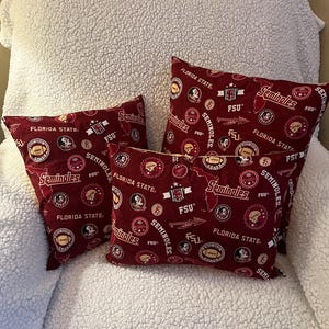 May include: Four decorative throw pillows with a maroon background and repeating patterns of the Florida State Seminoles logo, footballs, and the letters "FSU". The pillows are arranged on a white, textured surface.