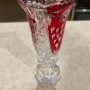 May include: A clear crystal vase with a red overlay. The vase has a flared top and a detailed, cut design. The base is round and also features a cut crystal design. The vase is approximately 8 inches tall.