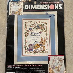 May include: A counted cross stitch kit by Dimensions, featuring a "Welcome Little One" design. The kit includes a framed image with a baby theme, featuring a mouse, duckling, and bunny reading a book. The frame size is 9" x 12" without mat.