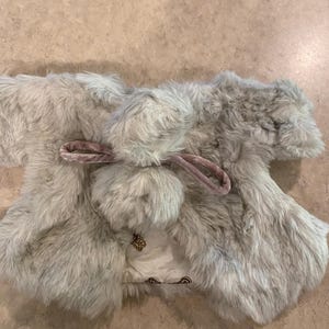 May include: A light gray faux fur vest with a matching pom-pom and pink velvet ties. The vest has a satin lining with a gold emblem. The vest is designed for a child.