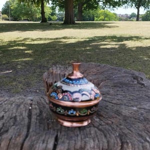 May include: A small, round, lidded container made of copper with a dark base and a cream-colored lid. The surface features intricate, hand-painted floral patterns in shades of blue, pink, and green, with a polished copper finial and base rim.