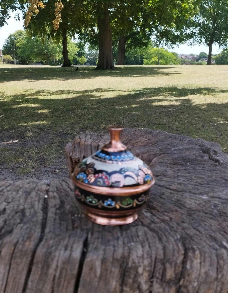 May include: A small, round, lidded container made of copper with a dark base and a cream-colored lid. The surface features intricate, hand-painted floral patterns in shades of blue, pink, and green, with a polished copper finial and base rim.