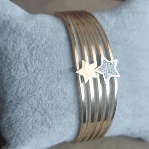 K&ouml;nnte beinhalten: Ein goldfarbenes Armband mit f&uuml;nf parallelen Reifen. Zwei sternf&ouml;rmige Anh&auml;nger sind an den Reifen befestigt; ein Stern ist aus glattem, goldfarbenem Metall, der andere ist mit kleinen, klaren, funkelnden Steinen besetzt.
