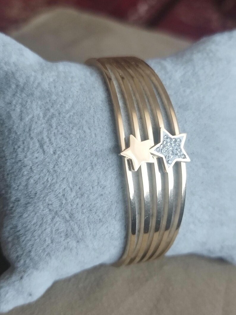 K&ouml;nnte beinhalten: Ein goldfarbenes Armband mit f&uuml;nf parallelen Reifen. Zwei sternf&ouml;rmige Anh&auml;nger sind an den Reifen befestigt; ein Stern ist aus glattem, goldfarbenem Metall, der andere ist mit kleinen, klaren, funkelnden Steinen besetzt.