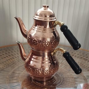 May include: A double-decker copper teapot featuring a polished metallic finish, two curved spouts, and two black cylindrical handles with gold-toned accents. The body of each pot is decorated with a repeating embossed floral pattern around the centre.