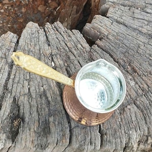 Pode incluir: Uma cafeteira de cobre com textura martelada e interior prateado. A cafeteira possui um cabo longo de cor clara e texturizado com um padr&atilde;o geom&eacute;trico. O recipiente tem uma abertura larga no topo e uma base arredondada para uso no fog&atilde;o.