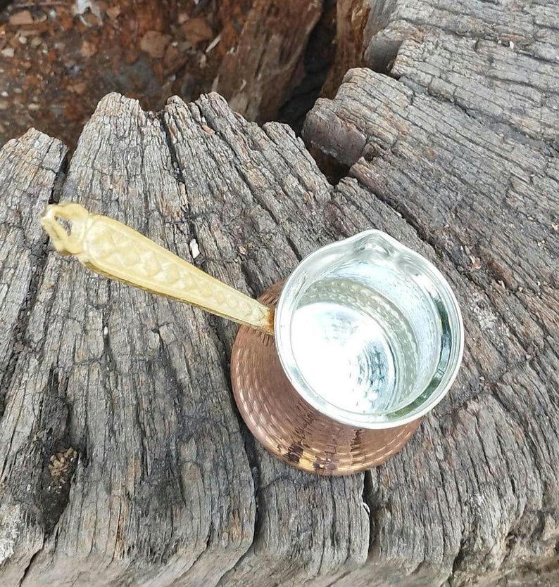 Pode incluir: Uma cafeteira de cobre com textura martelada e interior prateado. A cafeteira possui um cabo longo de cor clara e texturizado com um padr&atilde;o geom&eacute;trico. O recipiente tem uma abertura larga no topo e uma base arredondada para uso no fog&atilde;o.