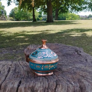 Peut inclure: Un petit r&eacute;cipient rond avec couvercle, fabriqu&eacute; en cuivre avec une finition bleu sarcelle. Le couvercle et la base pr&eacute;sentent des motifs floraux peints &agrave; la main dans des tons dor&eacute;s et cuivr&eacute;s, avec un bouton en cuivre poli sur le dessus.