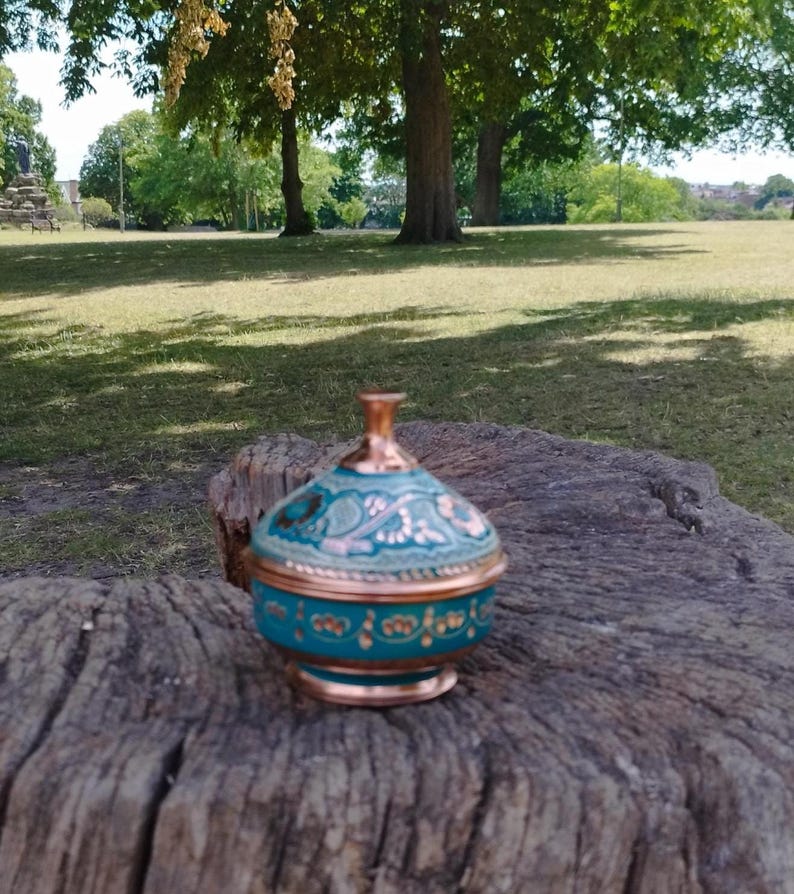 Peut inclure: Un petit r&eacute;cipient rond avec couvercle, fabriqu&eacute; en cuivre avec une finition bleu sarcelle. Le couvercle et la base pr&eacute;sentent des motifs floraux peints &agrave; la main dans des tons dor&eacute;s et cuivr&eacute;s, avec un bouton en cuivre poli sur le dessus.