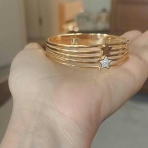 K&ouml;nnte beinhalten: Ein goldfarbener Klapparmreif mit vier parallelen B&auml;ndern. Das Design umfasst zwei sternf&ouml;rmige Akzente, einen schlichten goldfarbenen Stern und einen kleineren, mit kleinen klaren Steinen besetzten Stern auf der rechten Seite der B&auml;nder.