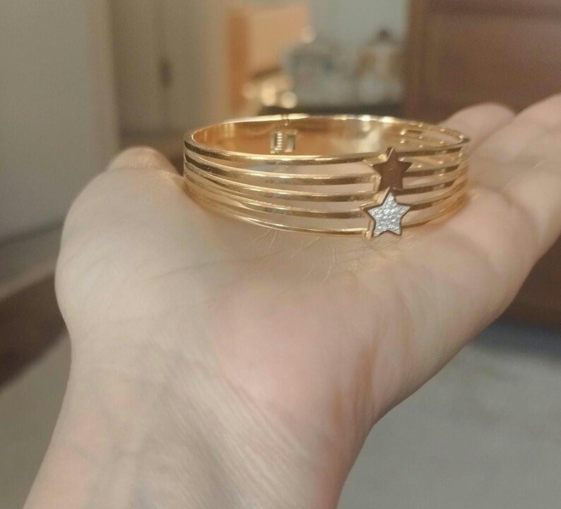 K&ouml;nnte beinhalten: Ein goldfarbener Klapparmreif mit vier parallelen B&auml;ndern. Das Design umfasst zwei sternf&ouml;rmige Akzente, einen schlichten goldfarbenen Stern und einen kleineren, mit kleinen klaren Steinen besetzten Stern auf der rechten Seite der B&auml;nder.