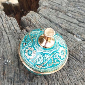 Peut inclure: Un r&eacute;cipient m&eacute;tallique rond avec un couvercle dot&eacute; d'une finition &eacute;maill&eacute;e turquoise vive et de motifs floraux et g&eacute;om&eacute;triques dor&eacute;s complexes. Le couvercle est surmont&eacute; d'un bouton cuivr&eacute; poli et la base est bord&eacute;e d'un liser&eacute; m&eacute;tallique.