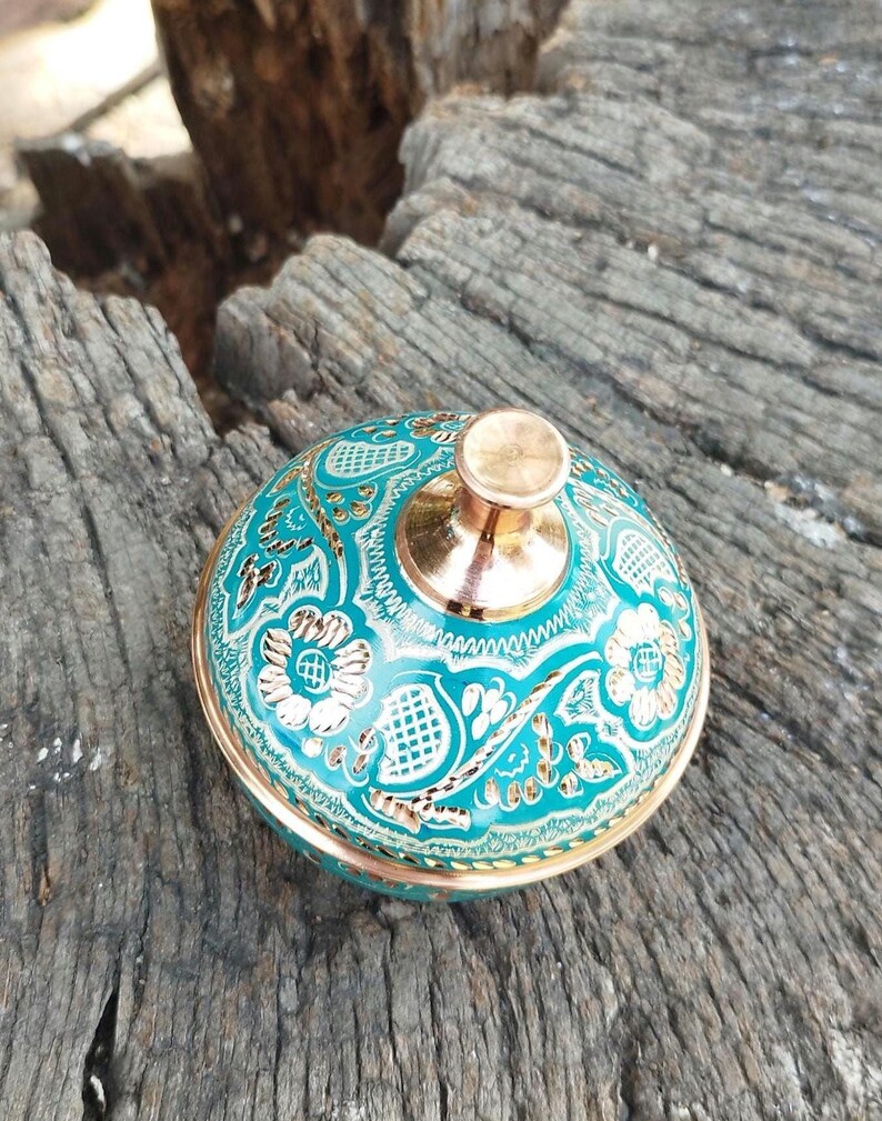 Peut inclure: Un r&eacute;cipient m&eacute;tallique rond avec un couvercle dot&eacute; d'une finition &eacute;maill&eacute;e turquoise vive et de motifs floraux et g&eacute;om&eacute;triques dor&eacute;s complexes. Le couvercle est surmont&eacute; d'un bouton cuivr&eacute; poli et la base est bord&eacute;e d'un liser&eacute; m&eacute;tallique.