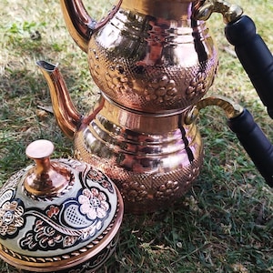 May include: A two-tiered copper teapot with a polished finish, featuring embossed floral patterns on the body, a curved spout, and black handles. Beside it sits a matching round copper bowl with a lid, decorated with black and white floral motifs.