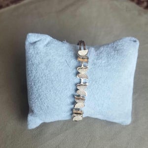 K&ouml;nnte beinhalten: Ein silberfarbenes Armband, das aus einer Reihe miteinander verbundener schmetterlingsf&ouml;rmiger Glieder besteht. Jedes Schmetterlingsglied hat eine texturierte, matte Oberfl&auml;che, die ein sich wiederholendes Muster entlang des Schmuckst&uuml;cks bildet.
