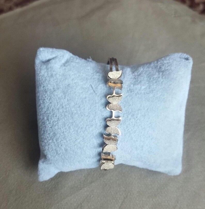 K&ouml;nnte beinhalten: Ein silberfarbenes Armband, das aus einer Reihe miteinander verbundener schmetterlingsf&ouml;rmiger Glieder besteht. Jedes Schmetterlingsglied hat eine texturierte, matte Oberfl&auml;che, die ein sich wiederholendes Muster entlang des Schmuckst&uuml;cks bildet.
