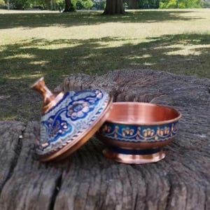 May include: A small copper bowl with a matching lid, featuring a vibrant blue and white floral enamel pattern. The bowl has a rounded base and a flared rim, while the lid includes a pointed handle, both decorated with intricate gold-toned details.