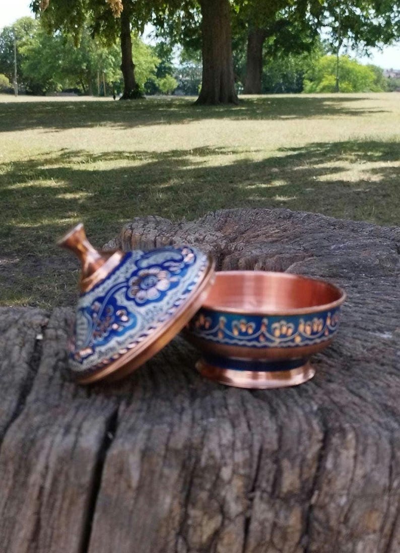 May include: A small copper bowl with a matching lid, featuring a vibrant blue and white floral enamel pattern. The bowl has a rounded base and a flared rim, while the lid includes a pointed handle, both decorated with intricate gold-toned details.