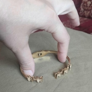 K&ouml;nnte beinhalten: Ein goldfarbenes Scharnierarmband mit einer Reihe kleiner, texturierter Schmetterlingsmotive entlang des Bandes. Das Armband verf&uuml;gt &uuml;ber einen sicheren Verschlussmechanismus und ein offenes Design mit einer detaillierten metallischen Oberfl&auml;che.