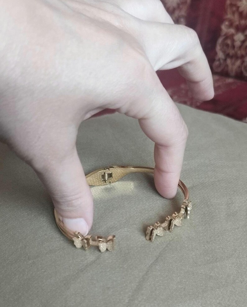 K&ouml;nnte beinhalten: Ein goldfarbenes Scharnierarmband mit einer Reihe kleiner, texturierter Schmetterlingsmotive entlang des Bandes. Das Armband verf&uuml;gt &uuml;ber einen sicheren Verschlussmechanismus und ein offenes Design mit einer detaillierten metallischen Oberfl&auml;che.