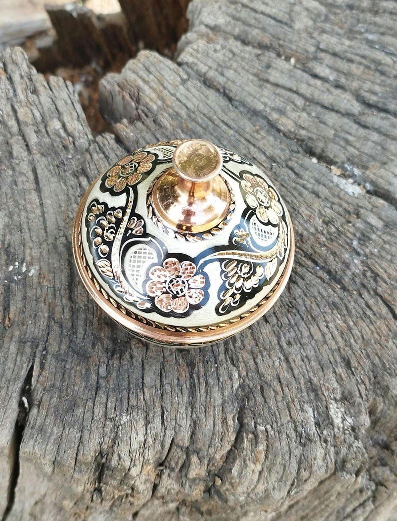May include: A round metal container lid featuring a polished copper-colored knob handle. The surface is decorated with a cream-colored background, black enamel accents, and intricate floral patterns etched in a metallic copper finish.